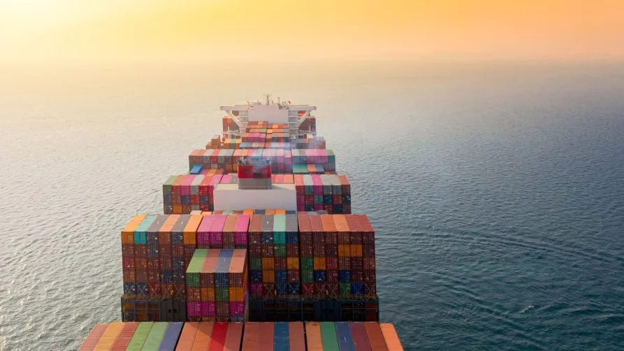 A large cargo ship loaded with colorful shipping containers sails through calm ocean waters under a golden sunset sky.