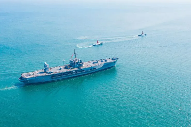 Aerial view of a military vessel navigating through calm, turquoise waters, accompanied by two smaller patrol boats. The ship appears to be an aircraft carrier or a naval command ship, moving steadily with the city skyline faintly visible in the background.