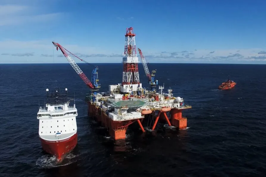 An offshore oil rig in the open sea, supported by large pillars and equipped with cranes and platforms. A supply vessel is approaching the rig, while another support boat is visible in the distance under a clear blue sky. AdobeStock