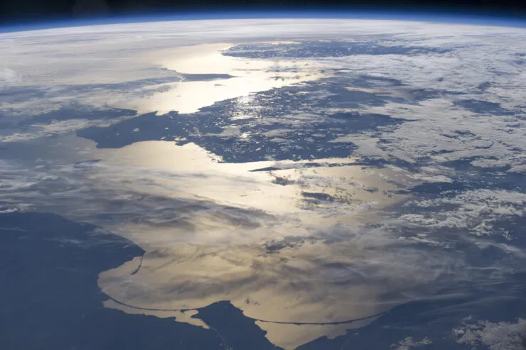 View of Earth from space, capturing a sunlit coastline with shimmering water reflecting the sunlight. Partially covered by scattered clouds, the landmass and ocean create a striking contrast, with the curvature of the Earth visible at the horizon. © Unseenlabs