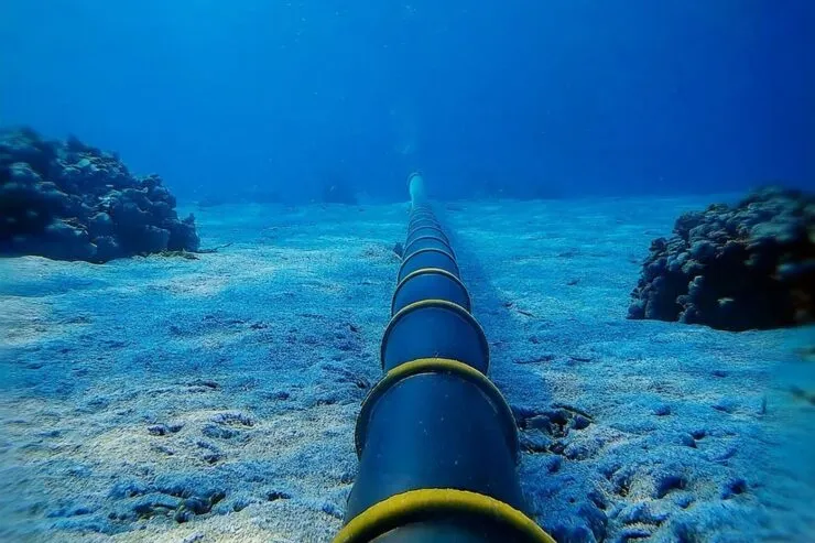 An underwater photograph of a submarine cable stretching across the ocean floor. The black cable, reinforced with yellow rings, extends into the distance, surrounded by a sandy seabed and coral formations. Sunlight filters through the clear blue water above. © Unseenlabs