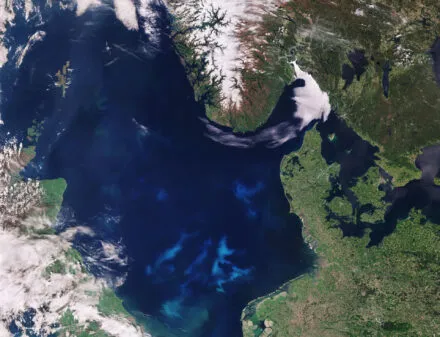 Satellite image of the North Sea and surrounding regions, including parts of the United Kingdom, Denmark, Norway, and Germany. The image shows swirling blue-green patterns in the water, indicating a phytoplankton bloom. Snow-covered mountains in Norway contrast with the lush green land areas, while clouds partially cover parts of the scene. Contains modified Copernicus Sentinel data (2017), processed by ESA