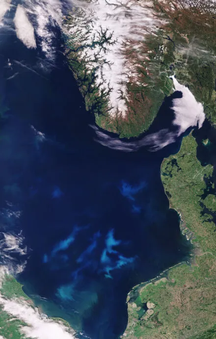 Satellite image of the North Sea and surrounding regions, including parts of the United Kingdom, Denmark, Norway, and Germany. The image shows swirling blue-green patterns in the water, indicating a phytoplankton bloom. Snow-covered mountains in Norway contrast with the lush green land areas, while clouds partially cover parts of the scene. Contains modified Copernicus Sentinel data (2017), processed by ESA