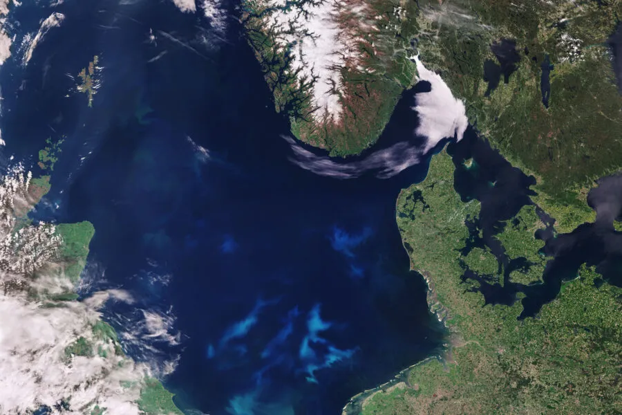 Satellite image of the North Sea and surrounding regions, including parts of the United Kingdom, Denmark, Norway, and Germany. The image shows swirling blue-green patterns in the water, indicating a phytoplankton bloom. Snow-covered mountains in Norway contrast with the lush green land areas, while clouds partially cover parts of the scene. Contains modified Copernicus Sentinel data (2017), processed by ESA