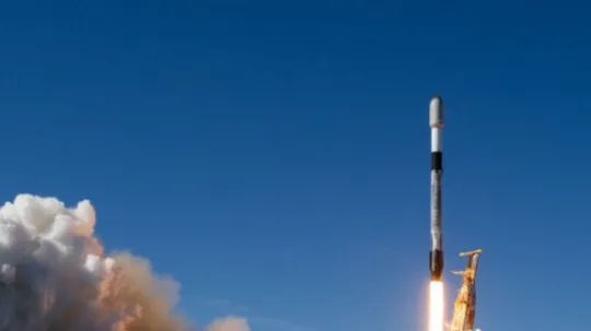 A rocket ascends into the sky, leaving behind a bright trail of fire and thick clouds of smoke. The launchpad structure beneath it is partially engulfed in the exhaust plume. The background features a clear blue sky and an ocean horizon, suggesting a coastal launch site. The rocket appears to be a modern, reusable launch vehicle, with visible markings and a sleek aerodynamic design.