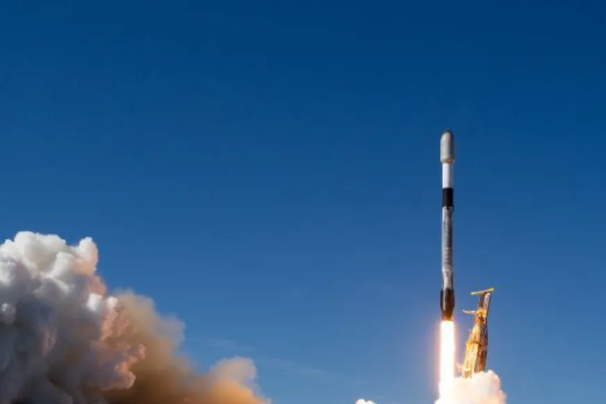 A rocket ascends into the sky, leaving behind a bright trail of fire and thick clouds of smoke. The launchpad structure beneath it is partially engulfed in the exhaust plume. The background features a clear blue sky and an ocean horizon, suggesting a coastal launch site. The rocket appears to be a modern, reusable launch vehicle, with visible markings and a sleek aerodynamic design.