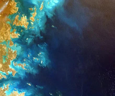 A satellite image capturing a coastal region with a striking contrast between land and sea. The shoreline is marked by intricate waterways, small islands, and vibrant turquoise waters blending into the deep blue ocean. The image showcases detailed topography, sediment flows, and ocean currents, highlighting the complexity of coastal ecosystems and maritime environments.