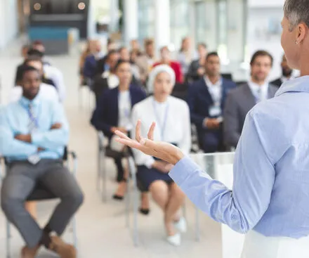 A professional woman with short gray hair, wearing a light blue blouse, stands at the front of a modern conference room, addressing a diverse audience. She gestures confidently as the attendees, dressed in business attire, listen attentively. The setting is bright and spacious, with large windows allowing natural light to illuminate the room.