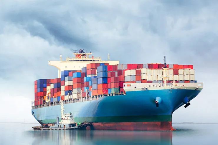 A large cargo ship loaded with colorful shipping containers is escorted by a smaller tugboat on calm waters under a cloudy sky. The image features the logos of the International Union of Marine Insurance (IUMI) and the ParisMAT conference, indicating a focus on maritime insurance and trade. The reflection of the ship and sky on the water enhances the serene yet powerful presence of the vessel.