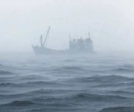 A fishing vessel emerges from dense fog over a rough and choppy sea. The scene is dominated by shades of gray and blue, with waves rolling in the foreground, creating a sense of isolation and uncertainty. The mist partially obscures the boat, giving it a ghostly and mysterious appearance, as if it is lost or navigating through harsh weather conditions.