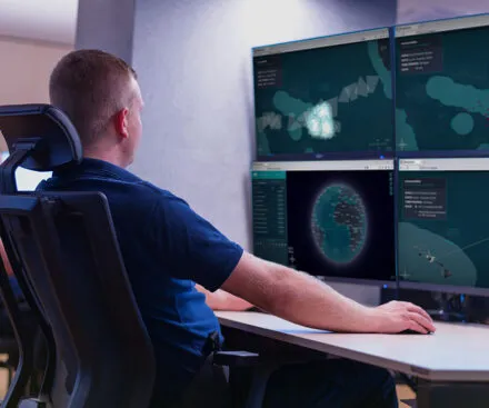 "A maritime surveillance officer monitors multiple data screens displaying real-time oceanic maps and vessel tracking information. The control center setup includes four large monitors with detailed visualizations of maritime activities, including ship routes, radar-like detection, and clusters of data points. The officer, dressed in a navy blue uniform, sits in a high-back ergonomic chair, attentively analyzing the information. The scene suggests a high-tech environment focused on maritime security, intelligence gathering, or vessel monitoring.