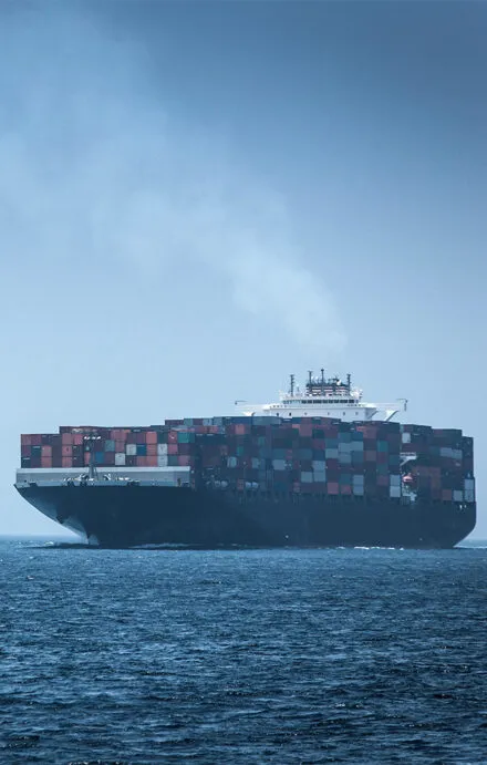 A large cargo ship loaded with multicolored shipping containers sails across the open ocean, emitting a trail of smoke from its exhaust stack. The vessel appears to be in transit, navigating through international waters under a hazy sky. The image highlights the scale of global trade and the environmental impact of maritime shipping.