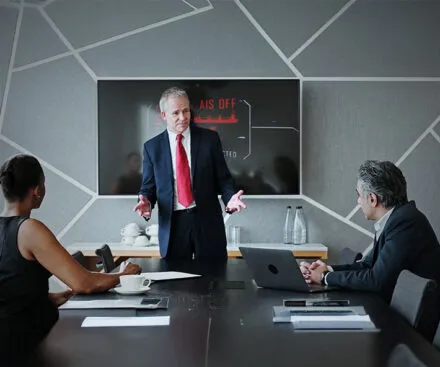 A business meeting in a modern conference room where a senior executive in a dark suit and red tie is presenting to two colleagues. A large screen behind him displays an alert reading "AIS OFF" with a graphic of a cargo ship, indicating a discussion about maritime security or risk management. The two attendees, a woman and a man in business attire, are engaged, with laptops and documents on the table. The setting suggests a high-level corporate discussion on shipping surveillance and risk mitigation.