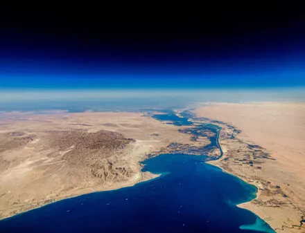 A high-altitude aerial view of the Suez Canal cutting through the arid desert landscape, connecting the Mediterranean Sea to the Red Sea. The canal serves as a crucial maritime trade route, with deep blue waters contrasting against the vast sandy terrain. Several ships are visible in the waterway, emphasizing the significance of global shipping and logistics. The sky transitions from a rich blue to a lighter hue near the horizon, adding depth to the image.
