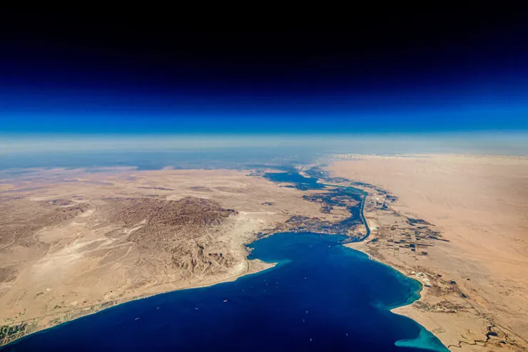 A high-altitude aerial view of the Suez Canal cutting through the arid desert landscape, connecting the Mediterranean Sea to the Red Sea. The canal serves as a crucial maritime trade route, with deep blue waters contrasting against the vast sandy terrain. Several ships are visible in the waterway, emphasizing the significance of global shipping and logistics. The sky transitions from a rich blue to a lighter hue near the horizon, adding depth to the image.