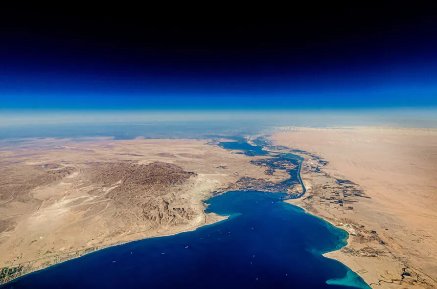A high-altitude aerial view of the Suez Canal cutting through the arid desert landscape, connecting the Mediterranean Sea to the Red Sea. The canal serves as a crucial maritime trade route, with deep blue waters contrasting against the vast sandy terrain. Several ships are visible in the waterway, emphasizing the significance of global shipping and logistics. The sky transitions from a rich blue to a lighter hue near the horizon, adding depth to the image.
