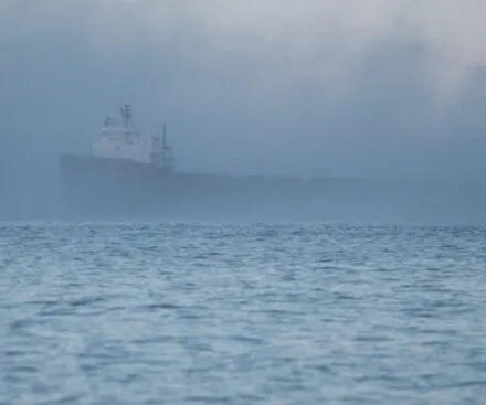 A large cargo ship emerges from dense fog over the open sea, its silhouette barely visible through the mist. The water in the foreground is calm, reflecting the eerie, muted tones of the sky. The scene evokes a sense of mystery and uncertainty, highlighting the challenges of maritime navigation in low-visibility conditions.