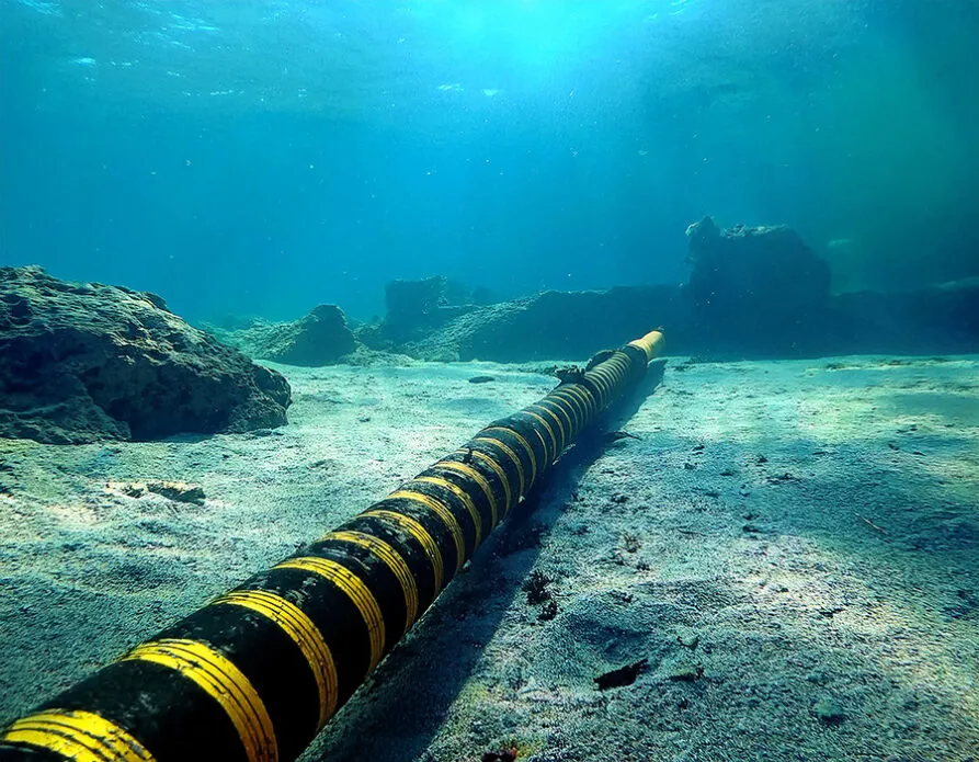 An underwater view of a large subsea cable with black and yellow protective coating, extending across the ocean floor. The cable is surrounded by a sandy seabed with scattered rocks and coral formations, illuminated by sunlight filtering through the clear blue water. This image highlights the critical role of subsea infrastructure in global telecommunications and energy transmission.