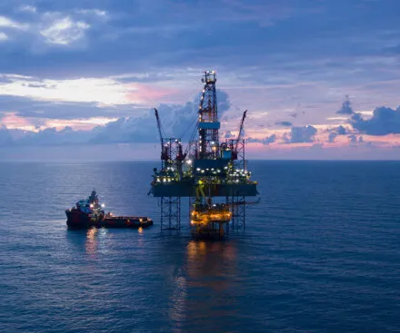 An offshore oil rig illuminated at dusk, standing in the middle of the ocean with a support vessel nearby. The rig's lights reflect on the calm waters, while a dramatic sky with scattered clouds adds depth to the background. The image captures the vastness of offshore operations and the strategic role of maritime infrastructure in energy production.