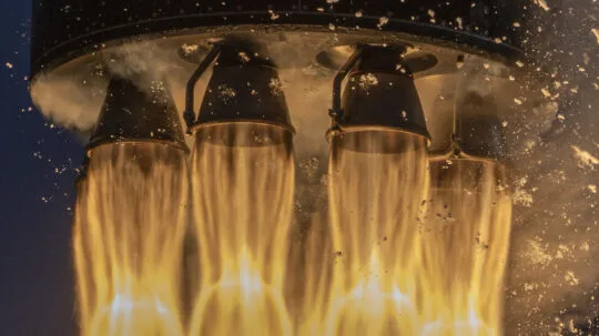 A close-up shot of a rocket's engine thrusters during launch, capturing the intense fiery exhaust plumes and glowing flames produced by the propulsion system. The image showcases four metallic nozzles expelling superheated gas, with bright orange and yellow flames contrasting against the dark background. Sparks and residual particles are visible around the engines, emphasizing the immense power and precision required for spaceflight. This dramatic image highlights the cutting-edge technology behind modern rocket launches and space exploration.