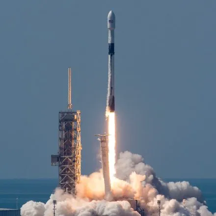 A Falcon 9 rocket lifts off from its launch pad, generating a bright plume of fire and thick clouds of smoke. The rocket, painted in black and white, ascends into a clear blue sky, with its first stage visibly separating below. The launch pad structure stands tall on the left, partially illuminated by the glow of the rocket's engines. The ocean is faintly visible in the background, adding to the dramatic scene of space exploration and technological advancement.