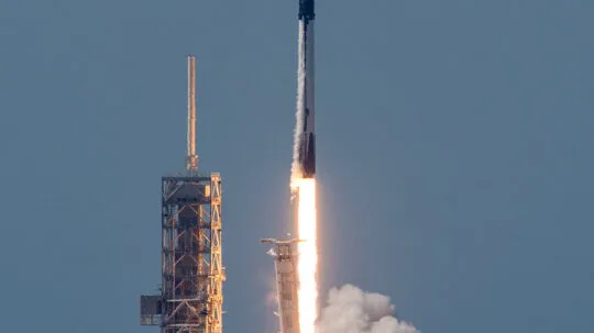 A Falcon 9 rocket lifts off from its launch pad, generating a bright plume of fire and thick clouds of smoke. The rocket, painted in black and white, ascends into a clear blue sky, with its first stage visibly separating below. The launch pad structure stands tall on the left, partially illuminated by the glow of the rocket's engines. The ocean is faintly visible in the background, adding to the dramatic scene of space exploration and technological advancement.