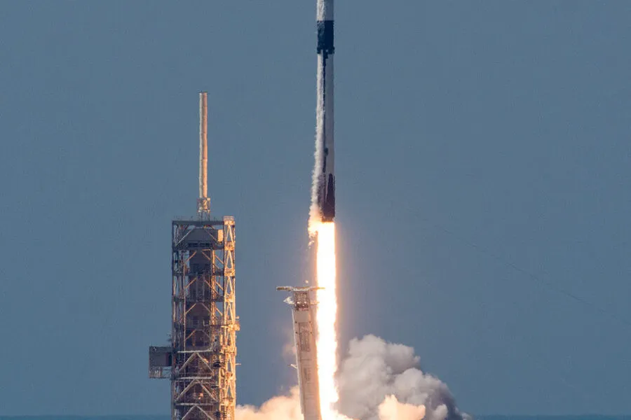 A Falcon 9 rocket lifts off from its launch pad, generating a bright plume of fire and thick clouds of smoke. The rocket, painted in black and white, ascends into a clear blue sky, with its first stage visibly separating below. The launch pad structure stands tall on the left, partially illuminated by the glow of the rocket's engines. The ocean is faintly visible in the background, adding to the dramatic scene of space exploration and technological advancement.