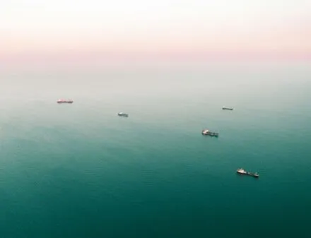 An aerial view of multiple cargo ships scattered across a vast, calm ocean with a soft pinkish horizon in the background. The vessels appear to be waiting or anchored in open waters.