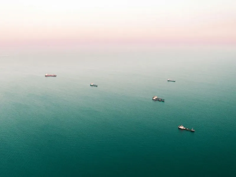 An aerial view of multiple cargo ships scattered across a vast, calm ocean with a soft pinkish horizon in the background. The vessels appear to be waiting or anchored in open waters.