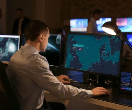 A professional analyst in a dimly lit operations center is focused on a large computer screen displaying a maritime surveillance interface. The map highlights a specific location with coordinates and ship tracking data. Multiple monitors in the background show additional maps, graphs, and analytical tools, while other team members in business attire are engaged in discussions. A desk lamp illuminates the workspace, emphasizing the importance of precision and real-time monitoring in maritime intelligence.