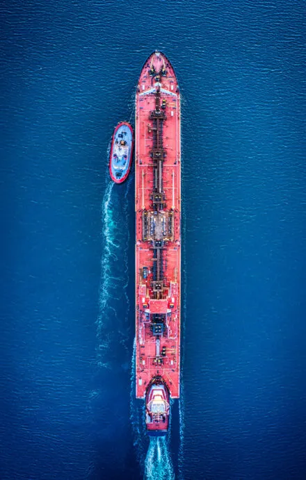 Aerial view of a large red tanker ship sailing through deep blue waters, accompanied by a smaller tugboat assisting in navigation. The image highlights maritime transportation, vessel operations, and the logistics of large-scale shipping.