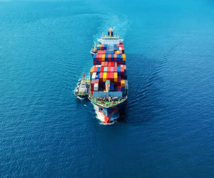 An aerial view of a large cargo ship sailing through the open ocean, fully loaded with multicolored shipping containers. Two smaller vessels flank the ship, possibly assisting with navigation or refueling. The deep blue sea stretches endlessly, emphasizing the scale of maritime logistics and global trade.