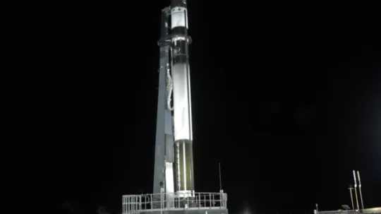A breathtaking long-exposure photograph captures the launch of a Rocket Lab vehicle carrying the BRO-1 satellite. The rocket's bright trail forms a perfect vertical beam of light piercing through the dark, cloudy night sky. Below, rolling green hills and the launchpad are faintly illuminated by the rocket's powerful glow. The scene conveys a sense of technological achievement and the awe-inspiring spectacle of space exploration.