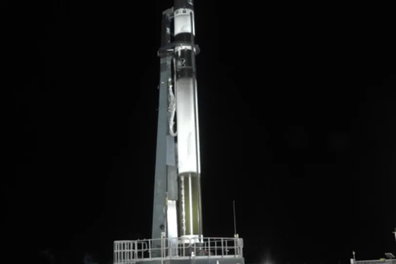 A breathtaking long-exposure photograph captures the launch of a Rocket Lab vehicle carrying the BRO-1 satellite. The rocket's bright trail forms a perfect vertical beam of light piercing through the dark, cloudy night sky. Below, rolling green hills and the launchpad are faintly illuminated by the rocket's powerful glow. The scene conveys a sense of technological achievement and the awe-inspiring spectacle of space exploration.