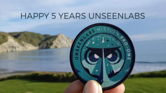 A hand holding an Unseenlabs Mission BRO-1 patch in front of a scenic coastal landscape with green hills and calm waters, with the text "HAPPY 5 YEARS UNSEENLABS" written above.