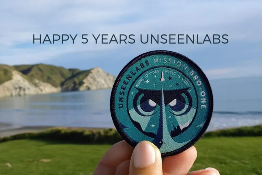 A hand holding an Unseenlabs Mission BRO-1 patch in front of a scenic coastal landscape with green hills and calm waters, with the text "HAPPY 5 YEARS UNSEENLABS" written above.