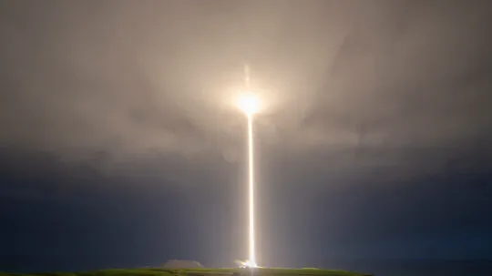 A bright rocket launch streaks vertically into a cloudy night sky, illuminating the landscape below with a powerful beam of light.