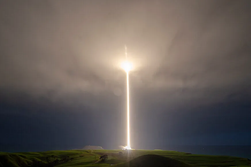 A bright rocket launch streaks vertically into a cloudy night sky, illuminating the landscape below with a powerful beam of light.
