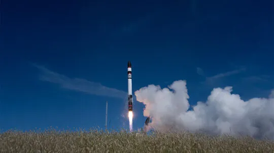 A rocket lifting off from a grassy field under a clear blue sky, leaving behind a plume of smoke and flame as it ascends.