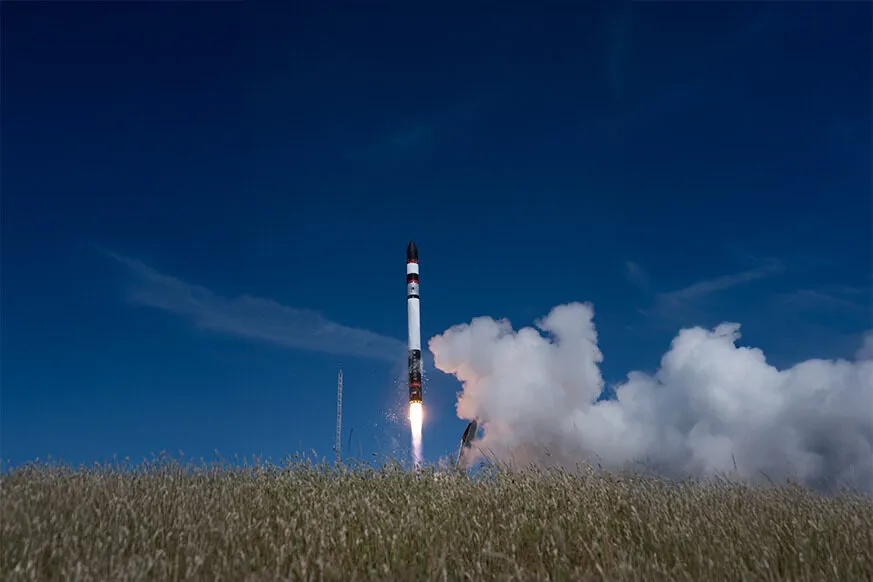 A rocket lifting off from a grassy field under a clear blue sky, leaving behind a plume of smoke and flame as it ascends.