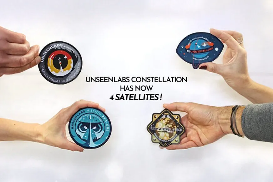 Four hands hold different mission patches representing Unseenlabs' satellites against a white background. Each patch has a distinct design, symbolizing a different mission. In the center, bold black text announces: "UNSEENLABS CONSTELLATION HAS NOW 4 SATELLITES!" The image visually highlights the growth of Unseenlabs' satellite constellation.
