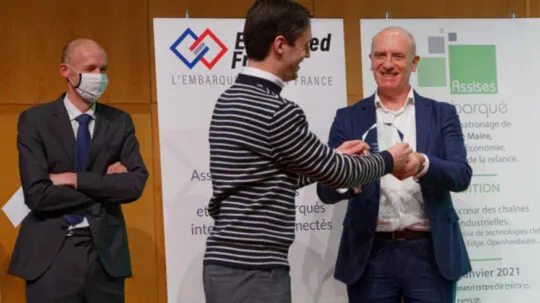 clément Galic in a striped sweater receives an award from another smiling man in a suit, while a third man, wearing a mask and standing with arms crossed, observes. Banners in the background display logos and text related to an embedded systems and IoT event.