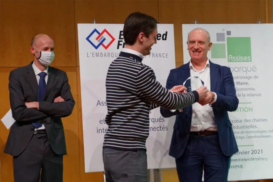 clément Galic in a striped sweater receives an award from another smiling man in a suit, while a third man, wearing a mask and standing with arms crossed, observes. Banners in the background display logos and text related to an embedded systems and IoT event.