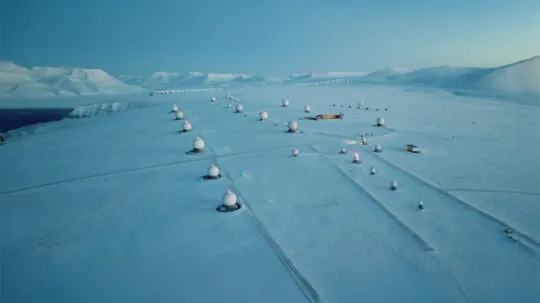 An aerial view of a snow-covered ground station in a remote Arctic landscape, with multiple white satellite antennas (radomes) spread across the icy terrain under a clear sky.