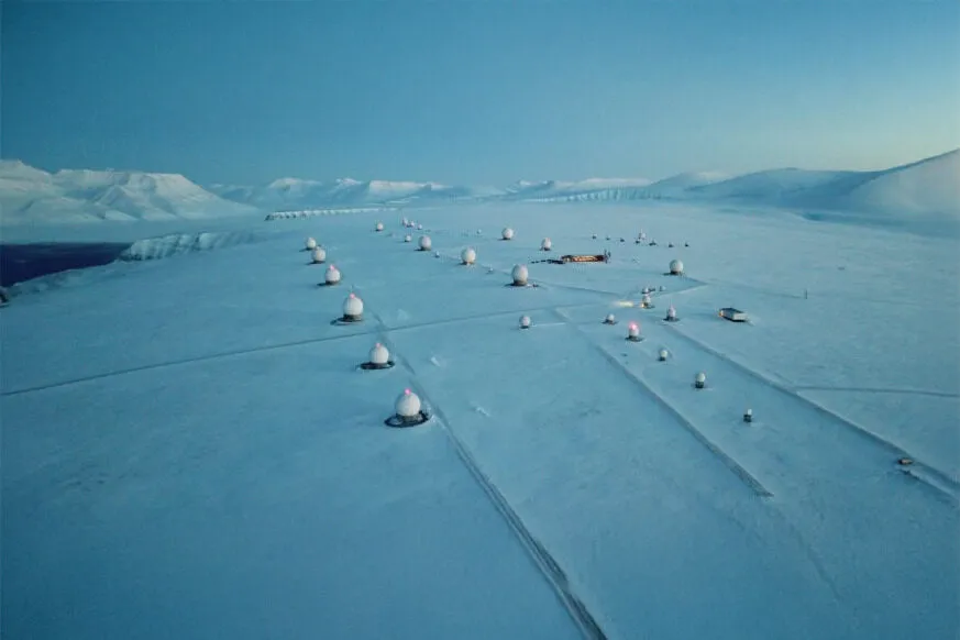 An aerial view of a snow-covered ground station in a remote Arctic landscape, with multiple white satellite antennas (radomes) spread across the icy terrain under a clear sky.