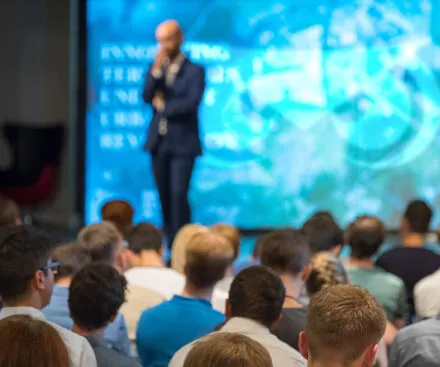 A speaker in a suit stands in front of a large, illuminated presentation screen displaying a futuristic digital interface. The audience, seen from behind, attentively listens to the talk in a professional conference setting. The image conveys an atmosphere of innovation, technology, and knowledge sharing.