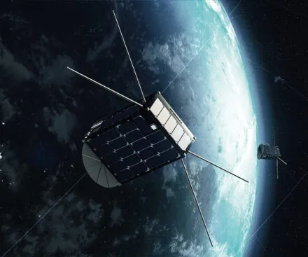A digital rendering of a satellite orbiting Earth in space. The satellite has a compact cubic structure with solar panels on its surfaces and long antennas extending outward. A second similar satellite is visible in the background, also orbiting the planet. The Earth appears partially illuminated, with a clear view of its atmosphere and cloud formations contrasting against the dark space.