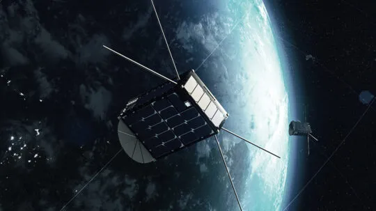 A digital rendering of a satellite orbiting Earth in space. The satellite has a compact cubic structure with solar panels on its surfaces and long antennas extending outward. A second similar satellite is visible in the background, also orbiting the planet. The Earth appears partially illuminated, with a clear view of its atmosphere and cloud formations contrasting against the dark space.