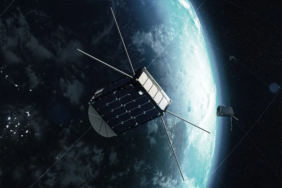 A digital rendering of a satellite orbiting Earth in space. The satellite has a compact cubic structure with solar panels on its surfaces and long antennas extending outward. A second similar satellite is visible in the background, also orbiting the planet. The Earth appears partially illuminated, with a clear view of its atmosphere and cloud formations contrasting against the dark space.