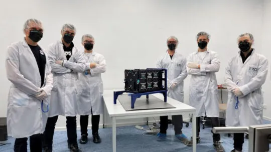 A team of six engineers and scientists wearing white lab coats, black face masks, hairnets, and gloves stand in a cleanroom around a small satellite mounted on a blue test stand. The satellite, covered in black panels with multiple connectors and components, is placed on a white table in a controlled environment. Some team members have their arms crossed, while others hold tools, signifying the final preparations before launch. The EXO logo is visible on their lab coats, indicating a collaboration with Exolaunch. The setting reflects a high-tech aerospace lab, emphasizing precision and innovation in satellite deployment.